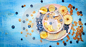 Berry greek yoghurt with frefh blueberries, banana and flakes in the pink bowl on the blue wooden table