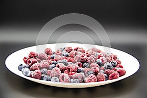 A Berry mix  from frozen raspberries and blueberries on the white plate. A Frozen Berries in black background.  A sweet background