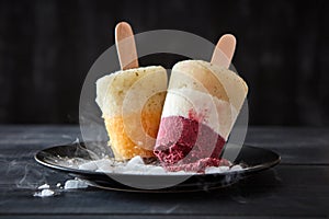 Berry sorbet lolly on a stick in a plate with slices of ice on a black wooden background. Dietary dessert