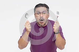 A bespectacled middle aged man looking awestruck while looking at something mouthwatering. Holding a fork and spoon hungry for a