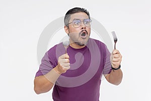 A bespectacled middle aged man looking in disbelief and shock while looking at something mouthwatering. Holding a fork and spoon