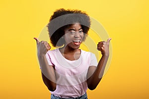 Best choice. Happy african american lady showing thumbs up gesture with both hands, posing on yellow studio wall