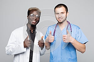 Best doctors. Two caucasian and afro american smiling doctors show a thumbs up while standing against gray background