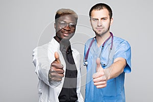 Best doctors. Two caucasian and afro american smiling doctors show a thumbs up while standing against gray background