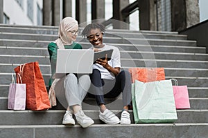 Best friends women making online purchases using a tablet and laptop sitting on steps of the mall