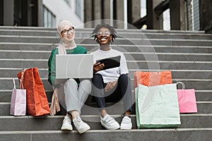 Best friends women making online purchases using a tablet and laptop sitting on steps of the mall