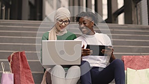 Best friends women making online purchases using a tablet and laptop sitting on steps of the mall