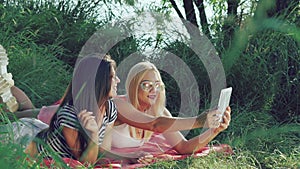 Best girl friends lying on grass and having fun taking a selfie with tablet