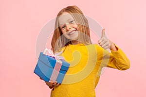 Best present! Portrait of lovely happy smiling little girl holding wrapped box and showing thumbs up, like gesture photo