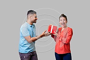 Best present for woman. Young handsome man in casual clothes giving gift box to pleasantly surprised woman. isolated on gray photo