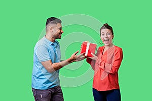 Best present for woman. Young handsome man in casual clothes giving gift box to pleasantly surprised woman. isolated on green