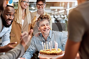 Best wishes. Young happy man blowing candles on cake while celebrating birthday among smiling mixed race business people