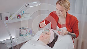 Beutician woman is applying clue mask on woman's face in beauty clinic.