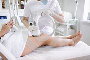 Beutiful bussines woman using phone while cosmetologist in white gloves performing radiofrequency lifting procedure on