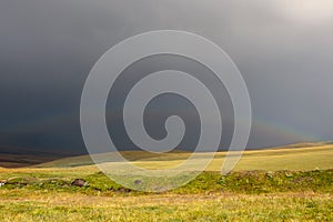 Beutiful rainbow above green meadow after heavy rain with rain clouds and mountains background. Summer season. Fresh green grass.