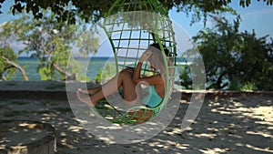 Beutiful young woman seats in the Hanging Chair, dreased in swimsewt , she relexing on the beack during her summer