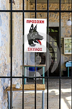 Beware of dog sign in Greek language on metal fence to outdoor rock entryway with tables - inside blurred behind bars