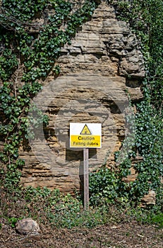 Beware of falling rock warning sign with triangle picture and alert message in yellow and black. Stood in front of cliff face.