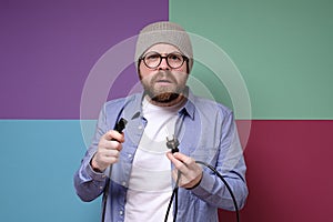 Bewildered bearded man, in round glasses and a hat, holds in hands an electric plug with a wire.