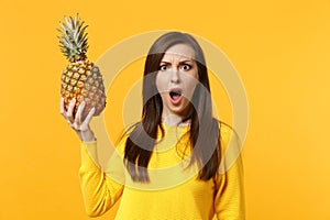 Bewildered irritated young woman in casual clothes hold, looking on fresh ripe pineapple fruit isolated on yellow orange