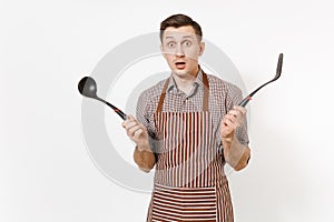 Bewildered man chef or waiter in striped brown apron, shirt holding black ladle or kitchen spoon, spatula isolated on