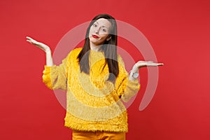 Bewildered perplexed young woman in yellow fur sweater spreading, pointing hands aside isolated on bright red wall