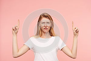 Bewildered young pretty redhead female with casual hairstyle showing up with forefingers and frowning confusedly her face while