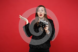 Bewildered young woman in black fur sweater spreading hand, holding retro vintage photo camera isolated on bright red
