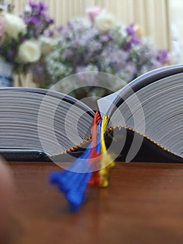 Bible book with yellow, red and blue ribbons marker on the wooden table with blur flowers bouquet background. Christianity.