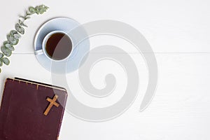 Bible and coffee cup and green leaves over the white wooden background.