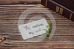 Biblical quote handwritten on lined paper with green plant and a closed holy bible book on wooden background