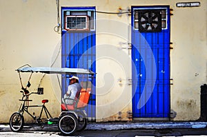 Bicitaxi driver parked up, infront of two blue doors, in the shade taking a rest.