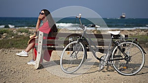 Bicycle leaning on bench outdoors with happy calm young woman sitting looking away on the left. Wide shot Mediterranean