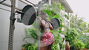 Biethnic woman performs barbell squats in rich outdoor home gym. Strength training exercise with fitness equipment