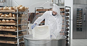 Big bakery industry baker in a special uniform preparing the dough add some more flour background worker moving the