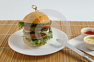 Big beef steak burger with vegetables and herbs on white plate and sauces on bamboo placemat