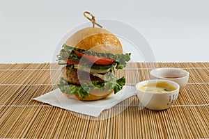 Big beef steak burger with vegetables and herbs on white plate and sauces on bamboo placemat