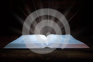 The big book of the Bible lies on a wooden table. In the dark. A light shines on the book from above. Light comes out of the book