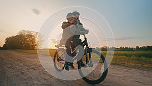 The big brother teaches the younger to ride a bike on a rural road at sunset.