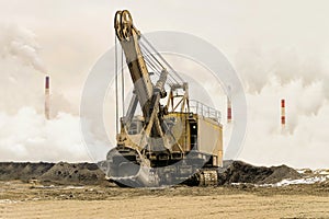 Big bucket mining excavator on a crawler track against a background of dense factory smog and fog with smoking chimneys. Heavy ind