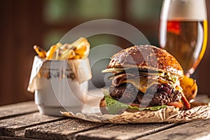 Big burger with beef, cheese, caramelized onion, lettuce, tomato, gherkin, all in a bun next to potato wedges and a glass of draft
