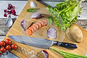 Big chef knife with healthy food - vegetables, onion, salad, potato placed on a cutting board with wood background top view