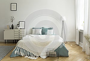 A big comfortable bed with pale sage green and white linen, pillows and blanket in a woman`s bright bedroom interior with windows