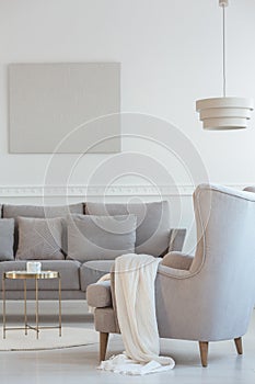 Big comfortable wing back armchair next to long grey scandinavian sofa with pillows in bright living room interior