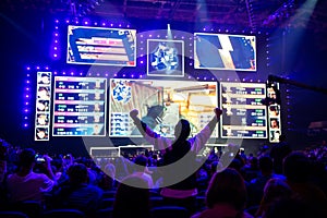 Big esports event. Video games fan on a tribune at tournament`s arena with hands raised. Cheering for his favorite team.