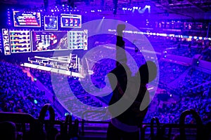 Big esports event. Video games fan on a tribune at tournament`s arena with hands raised. Cheering for his favorite team.