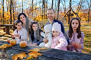 big family has picnic in autumn city park, children and parents sitting together at the table, with apples and yellow maple leaves