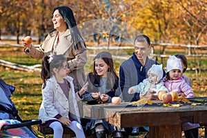 big family has picnic in autumn city park, children and parents sitting together at the table, with apples and yellow maple leaves