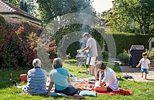 Big family having picnic and barbecue sitting on grass in garden at home at bright sunny day.