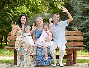 Big family sit on wooden bench in city park and waving, summer season, child, parent and grandmother, group of five people